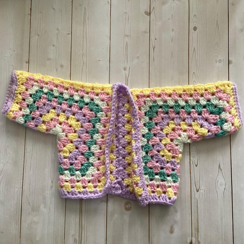 A crocheted cardigan with a white, pink, yellow, purple, pink, green, white, pink and yellow color pattern. The cardigan is open in the front and has a loose fit.