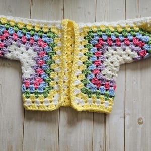 A crocheted cardigan with a white, purple, pink, blue, green, yellow, white and yellow color pattern. The cardigan is open in the front and has a loose fit.