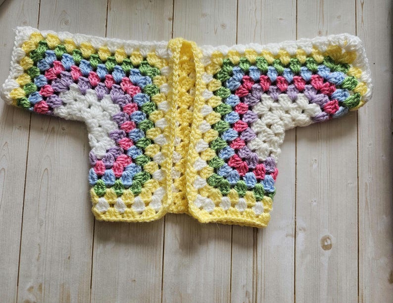 A crocheted cardigan with a white, purple, pink, blue, green, yellow, white and yellow color pattern. The cardigan is open in the front and has a loose fit.
