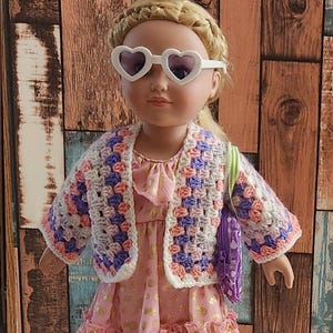 A doll wearing a crocheted cardigan with pink, purple, and white squares. The cardigan has a white trim. The doll is also wearing a pink dress with gold glitter and a purple purse with a green strap.
