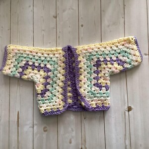 A pastel-colored, open-front crochet cardigan. The handmade garment features a granny square design in shades of yellow, purple, green, and pink. The edges are finished with a purple border. The cardigan is designed for a baby or toddler.
