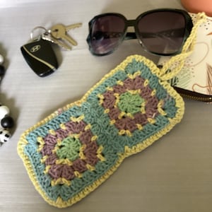Può includere: Un astuccio per occhiali all'uncinetto con un bordo giallo e un motivo a quadrato della nonna multicolore. L'astuccio è realizzato con un filo blu chiaro, verde e viola.