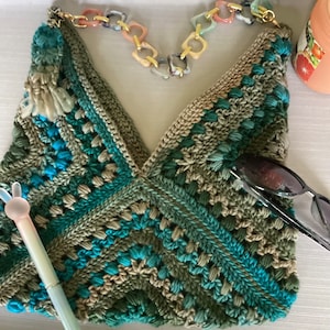 May include: A crocheted bag with a green, blue and beige pattern. The bag has a gold chain strap with colourful plastic links. The bag is lying on a white surface with a pair of sunglasses and a pink toy.
