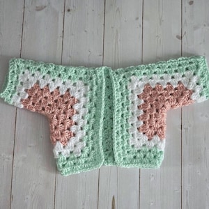 Pode incluir: Um cardigan de beb&ecirc; de croch&ecirc; com um padr&atilde;o de granny square. O cardigan apresenta um design de blocos de cores com quadrados verde-menta, brancos e p&ecirc;ssego. A pe&ccedil;a de roupa est&aacute; colocada sobre uma superf&iacute;cie de madeira clara.