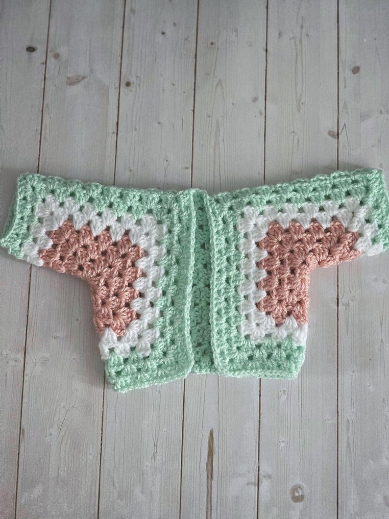 Pode incluir: Um cardigan de beb&ecirc; de croch&ecirc; com um padr&atilde;o de granny square. O cardigan apresenta um design de blocos de cores com quadrados verde-menta, brancos e p&ecirc;ssego. A pe&ccedil;a de roupa est&aacute; colocada sobre uma superf&iacute;cie de madeira clara.