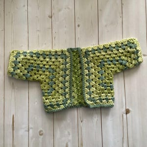 A crocheted cardigan with a light green and Dark green color pattern. The cardigan is open in the front and has a loose fit.