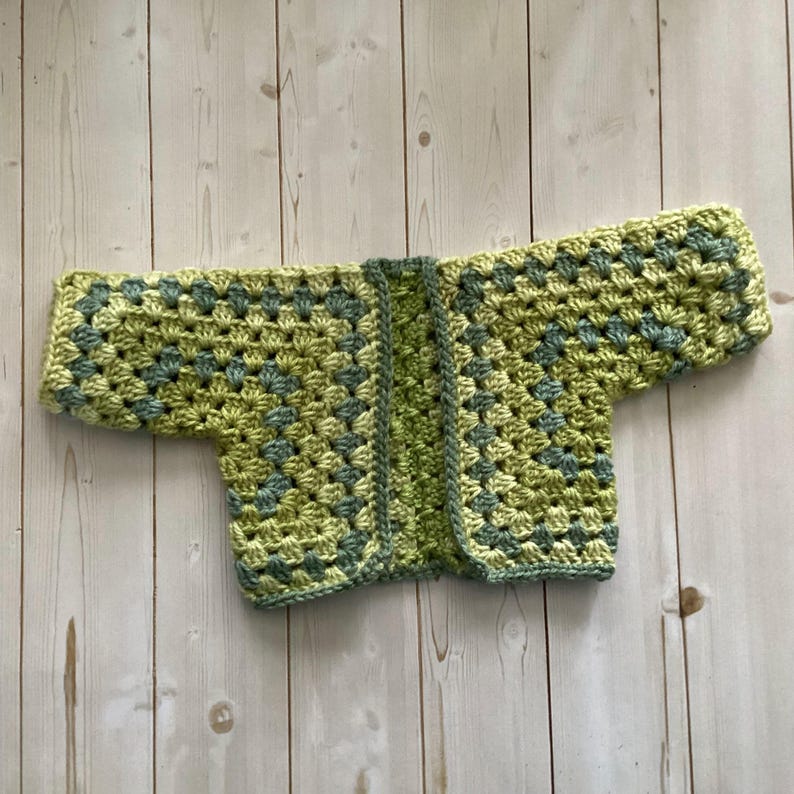A crocheted cardigan with a light green and Dark green color pattern. The cardigan is open in the front and has a loose fit.