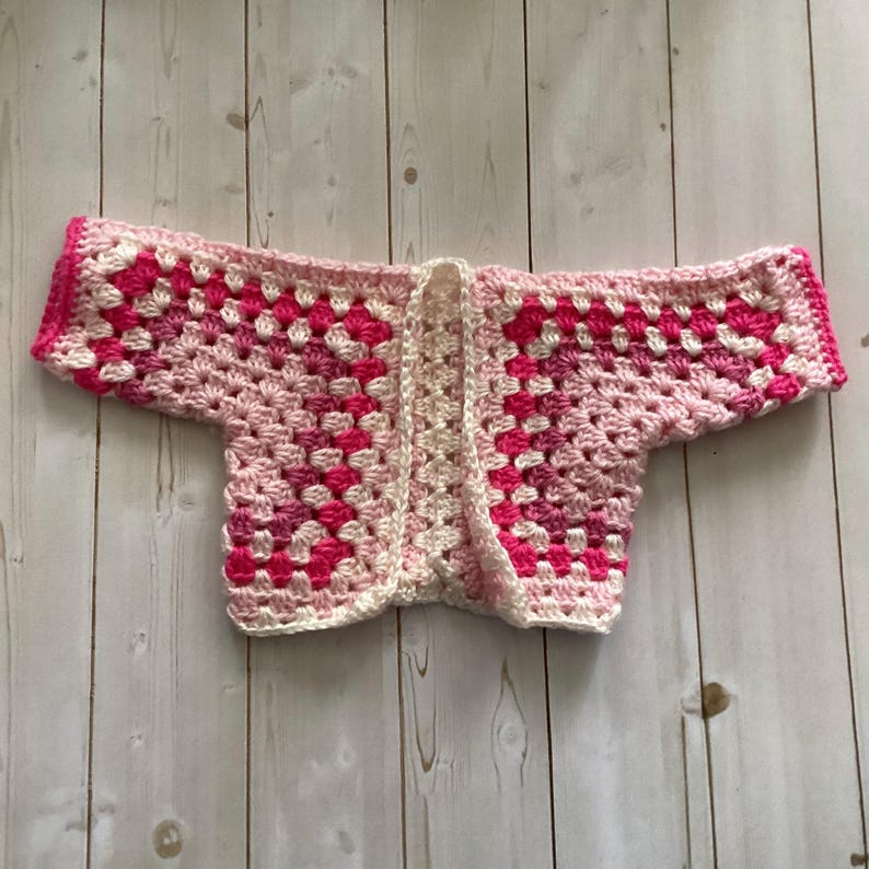 A crocheted cardigan with a black, brown, white, and blue color pattern. The cardigan is open in the front and has a loose fit.
