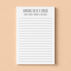 May include: A white rectangular notepad featuring black horizontal lines and the printed text HANGING ON BY A THREAD (OKAY, MAYBE A THREAD IS TOO THICK) in a simple sans-serif font at the top of the page, providing a clean space for writing notes.