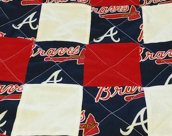 May include: A quilted blanket featuring a patchwork design with alternating squares of red, white, and navy blue fabric. The navy squares display the Atlanta Braves logo and script. The quilt is stitched with white thread.