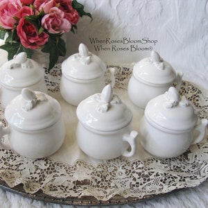 May include: Six white ceramic sugar bowls with lids, each featuring a handle and a decorative finial on top. The bowls are arranged on a silver tray with a lace doily, with a bouquet of pink roses in the background. Text on the image reads "When Roses Bloom Shop".