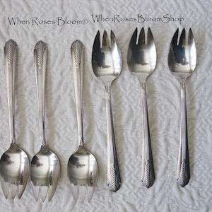 May include: Six silver serving spoons and forks are arranged on a white textured surface. The utensils have a decorative design on the handles and a fork-like end. The text "When Roses Bloom WhenRosesBloomShop" is at the top.