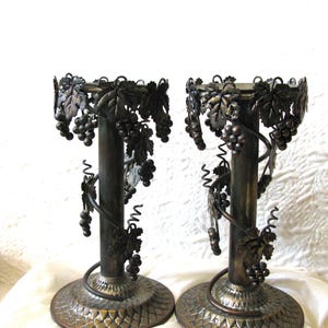 May include: A pair of dark bronze-toned metal candle holders featuring a cylindrical central pillar, a circular base with a textured scale pattern, and decorative metal grape clusters and vine leaves wrapping around the top and sides.
