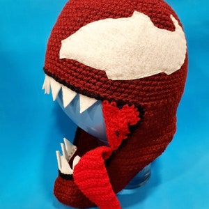 May include: A red and white crocheted hat with a large white face and a red tongue. The hat is designed to look like the Marvel Comics character Venom.