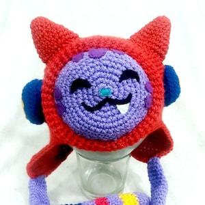 May include: A crocheted cat hat with a purple face, red ears, and a multi-colored tail. The hat has a blue and white eye and a pink nose.