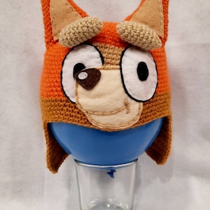 Peut inclure: Un bonnet en crochet orange et marron en forme de renard avec de grands yeux blancs et un nez marron. Le bonnet est posé sur un ballon bleu dans un verre transparent.