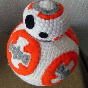 Może przedstawiać: Biała pluszowa zabawka w kształcie droida, wykonana na szydełku, z pomarańczowymi i szarymi detalami. Droid ma okrągłe ciało i mniejszą okrągłą głowę.