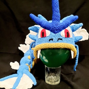 May include: Blue and white crocheted plush toy shaped like a dragon with red eyes. The toy is sitting on a glass with a green balloon inside.