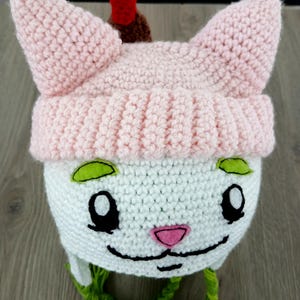 Puede incluir: Un gorro de ganchillo hecho a mano con forma de cara de gato. El gorro presenta orejas rosas, una cara blanca y una nariz verde y rosa. Tiene una correa verde y un detalle rojo y marrón en la parte superior.