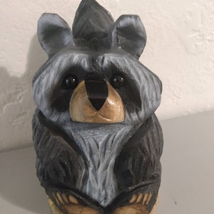 May include: A hand-carved wooden raccoon figurine. The raccoon has a black and gray face, ears, and body, with a light brown nose and paws. The figurine is sitting upright, with its paws resting on a wooden base. The carving has a textured appearance.