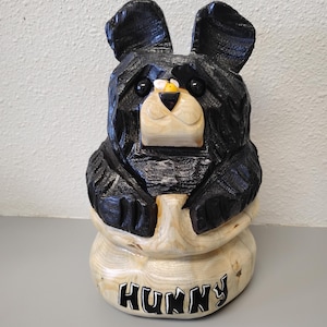 May include: A carved wooden bear figurine with black fur and a light-colored snout. The bear is holding a honey pot, with the word "HUNNY" in black letters on the base. The bear has a small yellow bee on its nose.