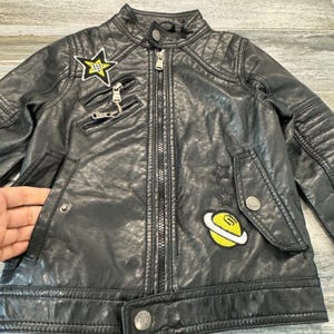 May include: Black faux leather jacket with a quilted design and a zipper closure. The jacket features decorative patches, including a yellow star, a planet, and stars. The jacket has a button closure at the waist and a small pocket.