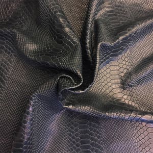 May include: A dark navy blue fabric featuring a textured, embossed snakeskin pattern. The material is draped in soft folds, highlighting the repeating scale-like design across the surface with subtle variations in light and shadow across the curves.