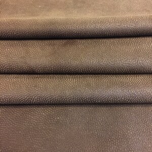 May include: Close-up of folded brown leather with a textured, bumpy surface. The leather has a dark, mottled appearance, with subtle variations in color and tone. The material appears to be high-quality, suitable for various crafting projects.