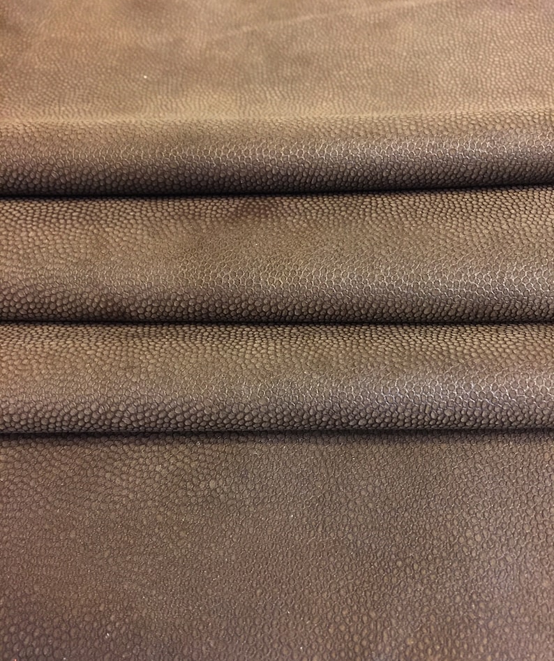 May include: Close-up of folded brown leather with a textured, bumpy surface. The leather has a dark, mottled appearance, with subtle variations in color and tone. The material appears to be high-quality, suitable for various crafting projects.