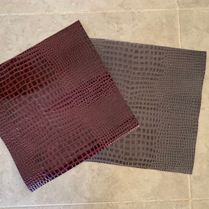 May include: Two sheets of faux leather with a crocodile skin texture. One sheet is a deep burgundy color, and the other is a dark gray. The sheets are rectangular and lie on a light gray tiled surface.