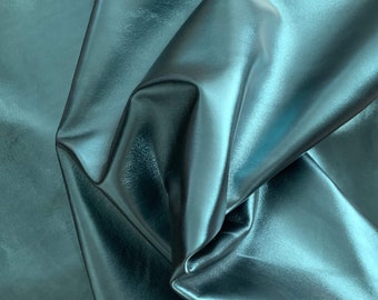 May include: Close-up of a teal-colored, metallic-looking fabric. The material is folded and draped, creating shadows and highlights that emphasize its texture and sheen. The fabric appears smooth and reflective, with a slight iridescent quality.