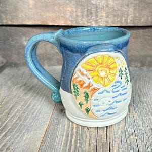 May include: A ceramic mug with a blue handle and rim, featuring a landscape design. The design includes a yellow sun, mountains, trees, and water. The mug is on a wooden surface.