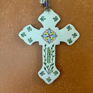 May include: White ceramic cross with a floral design. The cross features a central blue, yellow, and red floral pattern, green leaf accents, and a hanging loop. The cross is approximately 6 inches in height.