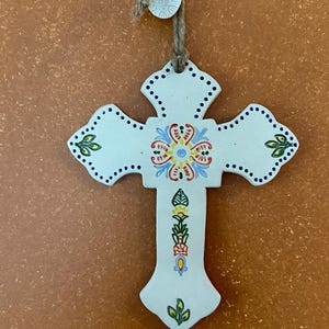 May include: A white ceramic cross with a floral design in the center, featuring red, blue, and yellow accents. The cross has a dotted border and is suspended by a natural twine cord. Decorative religious item.