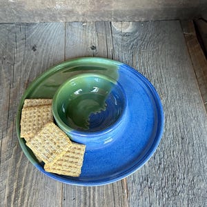May include: A handmade ceramic serving dish set with a green and blue color scheme. The set includes a large plate, a small bowl, and a few crackers. The bowl is nestled in the center of the plate. The set is on a wooden surface.