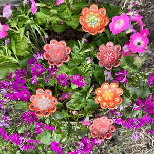 May include: A vibrant floral arrangement featuring a mix of pink, purple, and orange flowers. Decorative, flower-shaped objects are interspersed among the blooms. The arrangement is set against a backdrop of green foliage and dark soil.