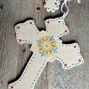 May include: Cream-colored ceramic cross with a floral design in the center, featuring blue, yellow, and red accents. The cross is adorned with blue dots and red flower-like details around the edges. A small, matching flower-shaped piece hangs above the cross.