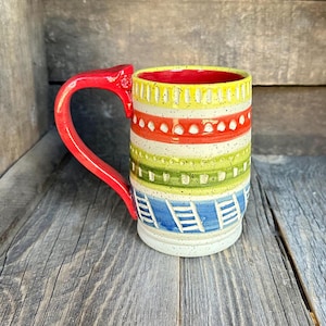 May include: A ceramic mug with a red handle and a colorful geometric pattern. The pattern includes stripes of red, yellow, green, and blue, with white accents.