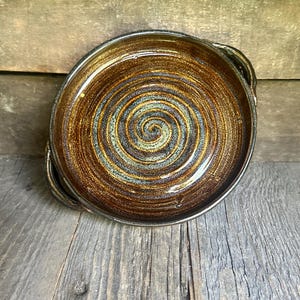 May include: A handmade ceramic plate with a spiral design. The plate features a brown and gold glaze with hints of blue and green. It has two small handles and is set on a wooden surface.