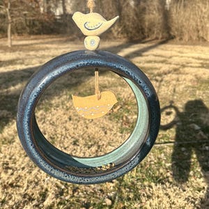 May include: A blue ceramic bird feeder with a round opening and a small bird figurine hanging from the top. The feeder is filled with birdseed and is hanging from a string.