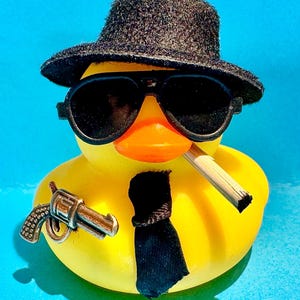 May include: A yellow rubber ducky wearing a black fedora, sunglasses, and a black tie. The ducky has a cigarette in its beak and a toy gun attached to its side. The background is a solid light blue.