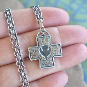 May include: A silver-toned cross pendant necklace. The cross has a textured surface with a dove emblem in the center. The necklace chain is a linked design.