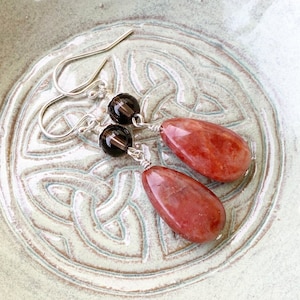 May include: A pair of dangle earrings with a teardrop-shaped red gemstone and a round brown gemstone. The earrings are made with silver wire and have silver hooks.