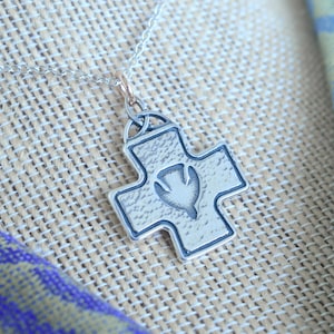 May include: A silver-colored pendant necklace featuring a cross design with a dove emblem in the center. The pendant hangs from a delicate silver chain. The background includes a woven fabric and a blue and green patterned cloth.