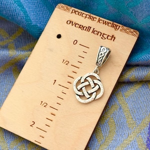 May include: A silver Celtic knot pendant charm with a bail, measuring 1 1/2 inches in length. The pendant is displayed on a wooden ruler with the text "peacefire jewelry overall length".