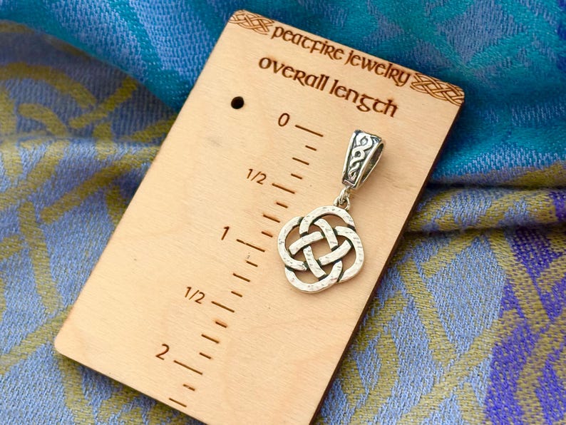 May include: A silver Celtic knot pendant charm with a bail, measuring 1 1/2 inches in length. The pendant is displayed on a wooden ruler with the text "peacefire jewelry overall length".