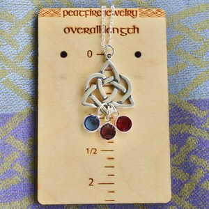 Puede incluir: Collar de plata con colgante de nudo celta y tres piedras preciosas colgantes en azul, rojo y morado. El collar se muestra en una regla de madera que mide 5 cm de largo.
