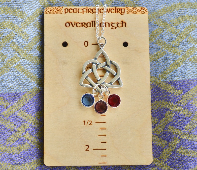 Puede incluir: Collar de plata con colgante de nudo celta y tres piedras preciosas colgantes en azul, rojo y morado. El collar se muestra en una regla de madera que mide 5 cm de largo.