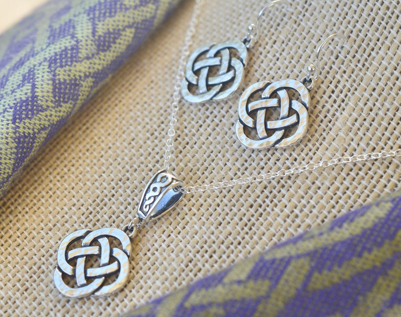 May include: A silver Celtic knot necklace and earrings set. The necklace features a pendant with a detailed knot design and a small charm with a Celtic knot. The earrings are also detailed with a Celtic knot design.