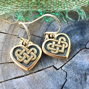 May include: Gold Celtic knot heart earrings with a dark green enamel finish. The earrings are on a wooden surface.